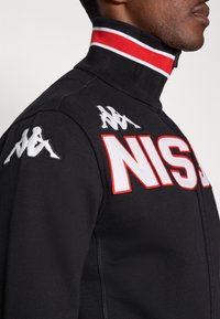 Svart jacka med dragkedja, röd- och vitrandig krage, stora bokstäver "NIS" och Kappa-logotyper i vit brodyr på axlarna.
