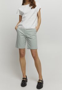 Hellgraue Shorts aus glattem Stoff mit Seitentaschen, kombiniert mit einem locker sitzenden weißen Oberteil. Schwarze Slipper mit goldenem Akzent.