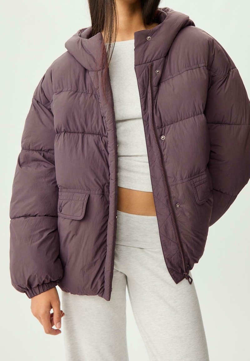 Femme portant une doudoune ample de couleur violet foncé sur un crop top gris et un pantalon de jogging gris, debout avec une main le long du corps.