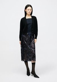 Cardigã preto, abotoado, sobre um top azul-marinho, combinado com uma saia preta transparente e com lantejoulas. Modelo usa meias pretas e sapatos de salto agudo.