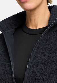 Schwarze Fleecejacke mit einer strukturierten Außenseite und glatter Innenseite. Verfügt über einen hohen Kragen und einen Reißverschluss. Wird über einem schwarzen Oberteil getragen.
