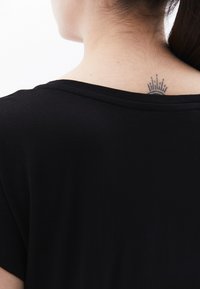 Mustahi, fupi wa nguo za shingo pana za rangi nyeusi. Kitambaa kinavyoonekana ni laini. Tattoo ya mviringo yenye mistari inayoangaza inaonekana kwenye mgongo wa juu.