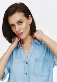 Jasnoniebieska, zapinana na guziki koszula z denimu z kołnierzykiem i krótkim rękawem; ma kieszeń na piersi i drewniane guziki.