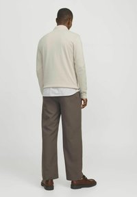 Šviesiai smėlinė ilgomis rankovėmis marškiniai su raukšlėta tekstūra, dėvimi virš baltų ir mėlynų dryžuotų marškinių, derinami su pilkais kelnėmis ir rudos spalvos batais.