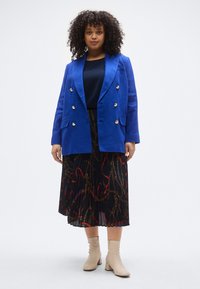 Kék kétsoros zakó arany gombokkal, fekete pliszírozott szoknya piros és arany láncmintával, valamint bézs bokacsizma.