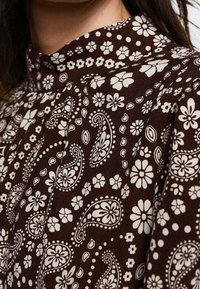 Brauner Stoff mit weißen Paisley- und Blumenmustern, ausgestattet mit einem hohen Kragen und gerafften Details, die eine strukturierte Oberfläche zeigen.