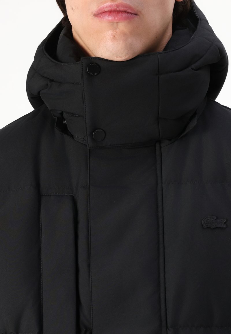 Veste matelassée noire avec un col montant, dotée de fermetures à pression et d'une surface texturée. Comprend un logo subtil sur la poitrine.