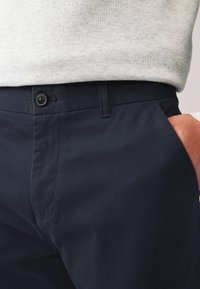 Navy plavi pamukasti chinos hlače s glatkom teksturom, ravnim prednjim dijelom, bočnim džepovima i jednim dugmetom na strateškoj završetku.
