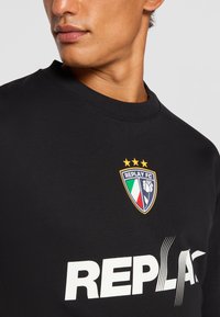 Black sweatshirt featuring a crew neckline, soft texture, large white "REPLAY" text, and a colorful logo with three stars above it.