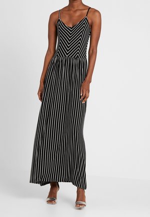 Femme portant une robe maxi noire sans manches avec des rayures blanches verticales et en chevrons, associée à des talons ouverts argentés.