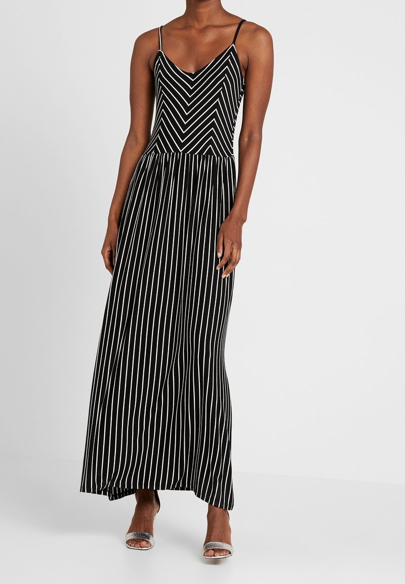 Femme portant une robe maxi noire sans manches avec des rayures blanches verticales et en chevrons, associée à des talons ouverts argentés.