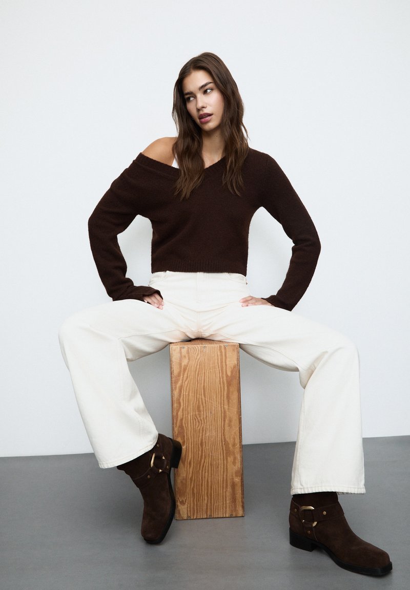 Pull en tricot marron avec un design décolleté, pantalon large crème, et bottines en daim marron avec une touche dorée, assis sur un bloc en bois.