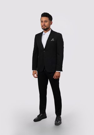 Mann mit kurzen schwarzen Haaren und Bart, der einen schwarzen Anzug, ein weißes Hemd, ein Einstecktuch und schwarze Schnürschuhe trägt, steht vor einem einfachen, hellgrauen Hintergrund.