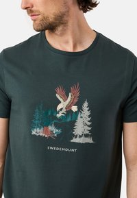 Mörkgrön bomullströja med en örn-grafik, träd och en bäck. "SWEDEMOUNT" text under designen. Rundhals, korta ärmar.