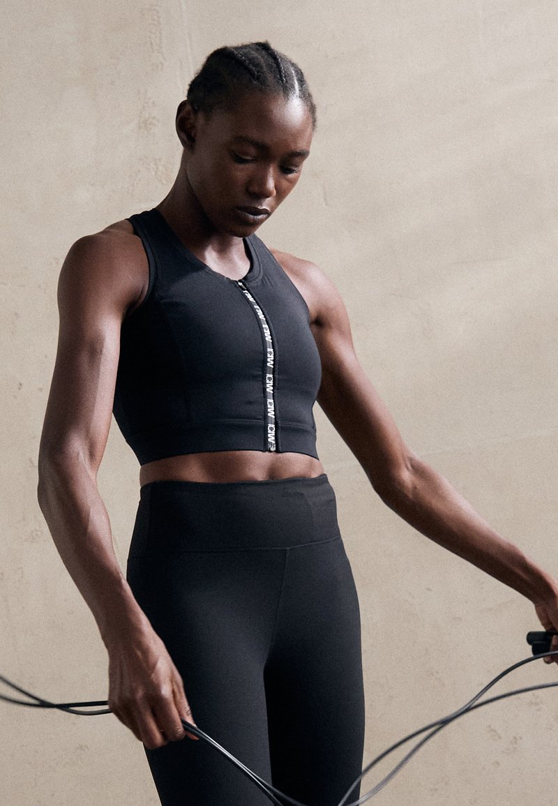 Top atlético preto de cropped com fecho na frente e detalhes de logotipo, combinado com leggings pretas de cintura alta; textura suave e justa.