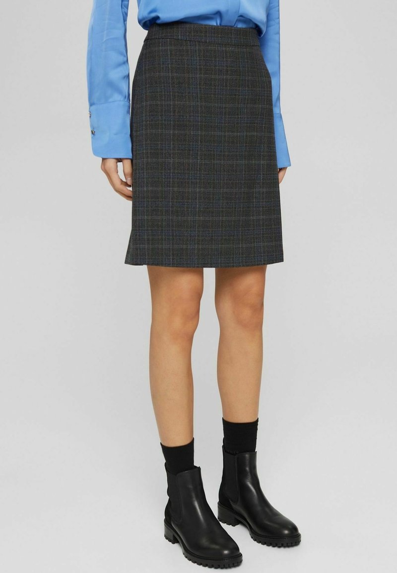 Femme portant une jupe grise à carreaux longueur genou, des bottines noires avec des semelles épaisses, des chaussettes noires et une chemise bleue à manches longues.