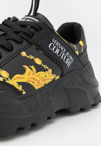 Crne atletske teniske s teksturiranim potplatom, žutim uzorkovanim detaljima i "VERSACE JEANS COUTURE" logotipom sa strane.