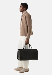 Schwarze Leder-Reisetasche mit zwei Griffen, rechteckiger Form und minimalistischem Design, gehalten von einer Person in beiger Jacke und weißer Hose.