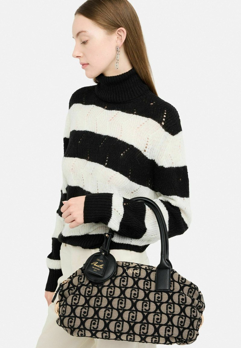 Sac en toile marron avec un motif de logo noir, poignées en cuir noir et quincaillerie dorée ; le modèle porte un pull rayé noir et blanc.