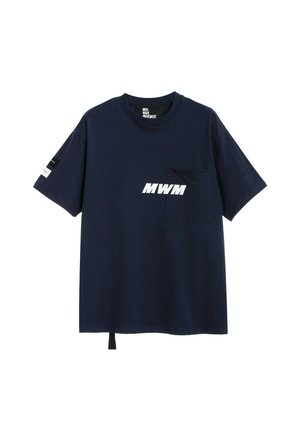 Joven con rastas vestido con una camiseta azul marino MWM, pantalones cortos blancos, calcetines blancos y zapatos beige con textura, sin cordones, sobre un fondo blanco liso.