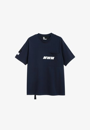 Joven con rastas vestido con una camiseta azul marino MWM, pantalones cortos blancos, calcetines blancos y zapatos beige con textura, sin cordones, sobre un fondo blanco liso.