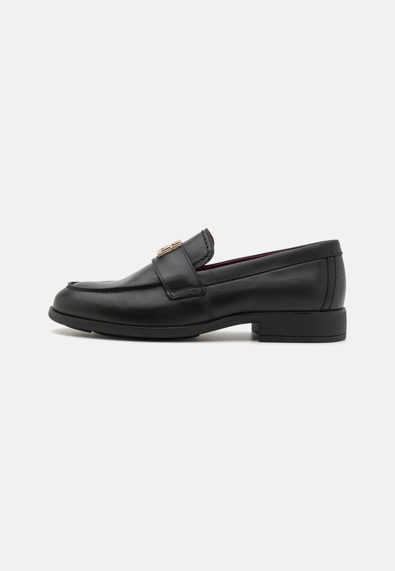 Mocassin en cuir noir avec un bout arrondi, un accent métallique décoratif à l'avant, et un petit talon carré. Texture lisse, design classique.