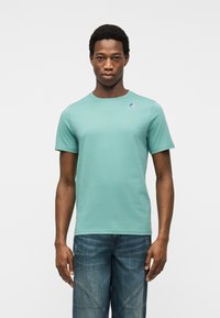 Νεαρός άνδρας που φοράει απλό ανοιχτό μπλε-πράσινο t-shirt με κοντά μανίκια και ένα μικρό λογότυπο στο στήθος, καθώς και μπλε τζιν, στέκεται μπροστά σε λευκό φόντο.