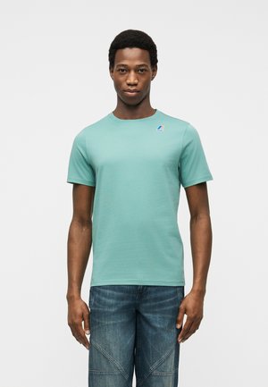 Mladý muž v jednoduchém světle tyrkysovém tričku s krátkými rukávy a malým logem na hrudi a modrých džínách stojí před bílým pozadím.