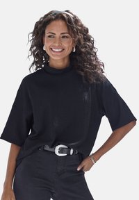 Schwarzes, strukturiertes Kurzarmoberteil mit hohem Kragen, getragen mit einem schwarzen Gürtel mit silberner Schnalle, kombiniert mit dunklen Hosen.