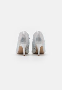 Un paio di scarpe con tacco alto argentate con decorazioni in strass, viste da dietro su uno sfondo bianco.