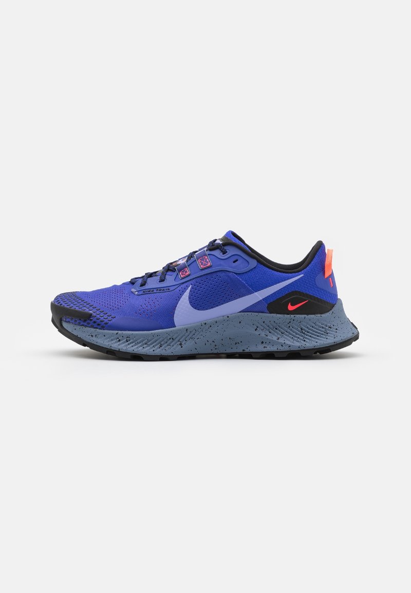 Chaussure de sport bleue avec une semelle grise mouchetée, des accents noirs et orange au talon, et un grand logo Nike en forme de swoosh bleu clair sur le côté.