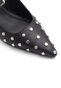 Chaussure en cuir noir avec un bout pointu, présentant de grandes bosses en argent sur les côtés pour la texture et les détails. Fermeture par boucle au niveau de la cheville.