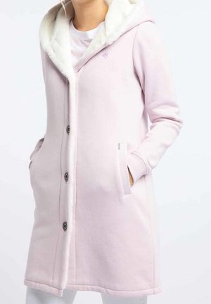 Femme portant un manteau rose clair mi-long aux genoux avec des boutons sur le devant, des poches latérales et une capuche doublée en polaire blanche sur une chemise blanche.