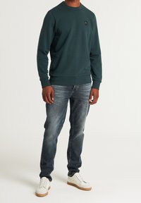 Mörkgrön huvtröja med rundad hals, ribbade ärmslut och nederkant. Kombinerad med bleka blå jeans och vita sneakers med tan färgade sulor. Svart logotypdetalj.