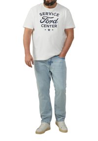 Man in een wit "Service Ford Center" t-shirt, lichtblauwe jeans en witte sneakers, staande met één hand in zijn zak.