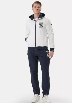 Uomo in piedi che indossa una giacca bianca con cerniera e cappuccio e pantaloni blu navy, entrambi con toppe ricamate Frecce Tricolori 61 Italia.