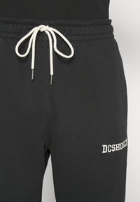 Zwarte joggingsbroek met witte trekkoord. Bevat logo tekst "DCSHOEC" in het wit op de rechteronderkant. Zacht materiaal en elastische tailleband.