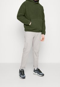 Felpa con cappuccio in pile verde scuro con tasca frontale, abbinata a pantaloni beige chiaro e sneaker grigie con dettagli neri e inserti in mesh.