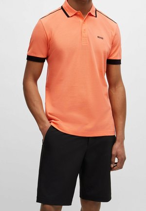 Mann trägt ein korallenfarbenes Polohemd mit schwarzem Rand und schwarze Shorts, steht mit teilweise in den Taschen befindlichen Händen vor einem einfarbigen Hintergrund.