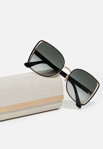 Černé sluneční brýle ve tvaru čtverce s tmavými skly, se zlatými akcenty, odpočívající na strukturovaném bílém pouzdře s béžovým páskem označeným "JIMMY CHOO."