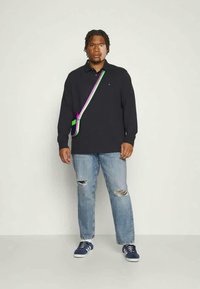 Marineblauw poloshirt met lange mouwen en een kraag, gecombineerd met vervaagde blauwe ripped jeans. Bevat een multikleurige schoudertas en marineblauwe sneakers.
