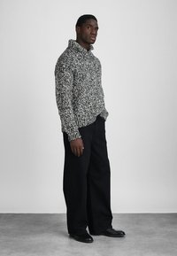 Άνδρας που φορά μαύρο και άσπρο πλεκτό πουλόβερ με ψηλό γιακά, μαύρο παντελόνι φαρδύ και μαύρα παπούτσια, στέκεται σε γκρι πάτωμα.