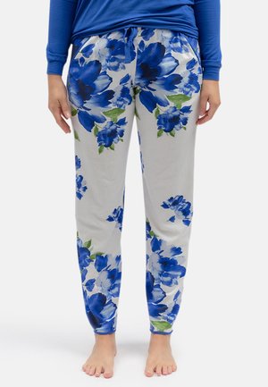 Persoon draagt witte broek met grote blauwe bloemenprint en groene bladeren, blootsvoets, staand tegen een effen achtergrond.