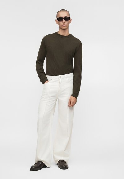 Άνδρας που φοράει μαύρα γυαλιά ηλίου, σκούρο πράσινο μακρυμάνικο πουκάμισο, λευκό παντελόνι με φαρδιά πόδια και μαύρα παπούτσια, όρθιος με το ένα χέρι στην τσέπη.