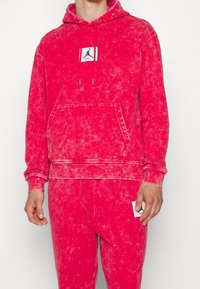 Červená batikovaná mikina s kapsou kangaroo, nastavitelným šňůrkovým uzávěrem a logo nášivkou. Měkká struktura s žebrovanými manžetami a lemem. Kombinováno s odpovídajícími kalhotami.