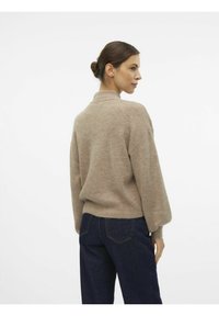 Beige stickad tröja med hög krage och ballongärmar, matchad med mörkblå jeans. Tyget ser mjukt ut med en subtil textur.