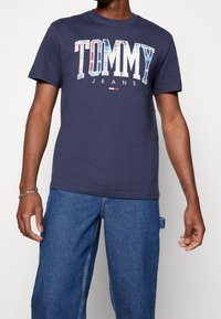 Marinblå bomulls-T-shirt med "TOMMY" i flerfärgad design, korta ärmar, rundad halsringning och slim fit. Kombinerad med denimbyxor.