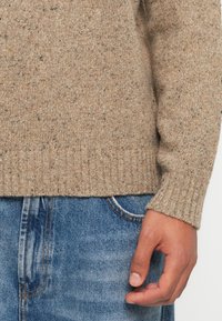 Villapaita vaaleanruskeana, jossa on pilkkuja, varustettuna ribbikäänteellä ja helmalla, yhdistettynä sinisiin farkkuihin. Pintakuvio on pehmeä ja lämmin.