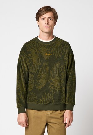 Jeune homme portant un sweat en polaire à motifs vert olive avec le logo "FAGUO", assorti à un pantalon beige, debout devant un fond uni.