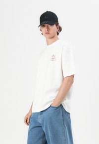 White cotton t-shirt featuring a small, colorful cup graphic on the chest. Paired with light blue denim shorts and a black cap.